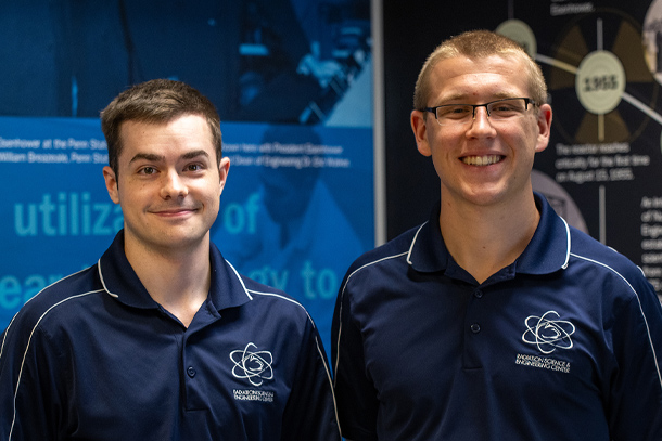 Two students wearing blue polos pose indoors behind a blue, black, and green backdrop. 